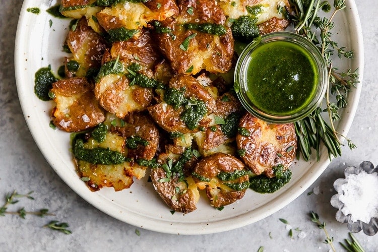 Overhead view several crispy smashed potatoes on stone plate drizzled with chimichurri sauce.