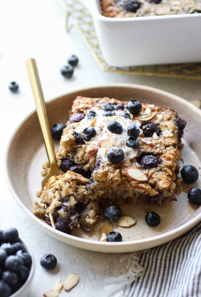 Blueberry Baked Oatmeal topped with whipped topping, drizzled with maple syrup on a small tan plate with a gold fork holding bite.
