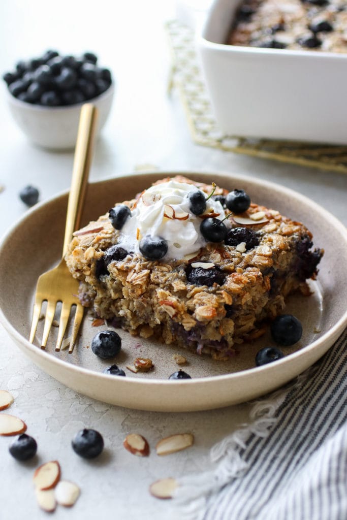 Healthy Blueberry Baked Oatmeal topped with whipped topping on a small tan plate with a gold fork resting on the plate.