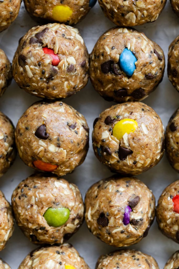A close up shot of monster cookie peanut butter balls lined up on a piece of parchment paper with colorful candy-coated chocolate pieces in each ball.
