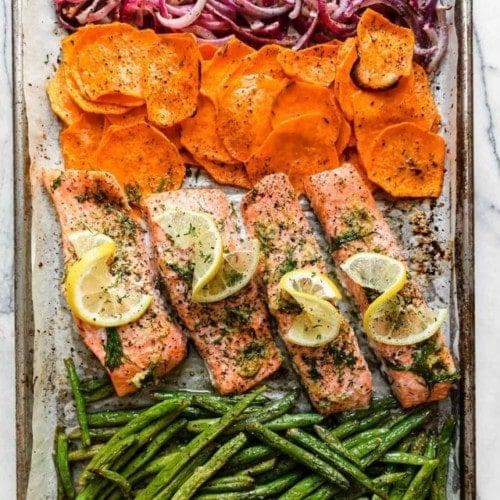 Overhead view of sheet pan filled with salmon fillets, sweet potato rounds, long fresh green beans, and red onions sliced.